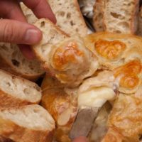 A hand holding a slice of bread, above a baked gruyere, using a knife to put cheese on the bread.