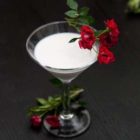 A cocktail in a martini glass, garnished with red spray roses on a black table.