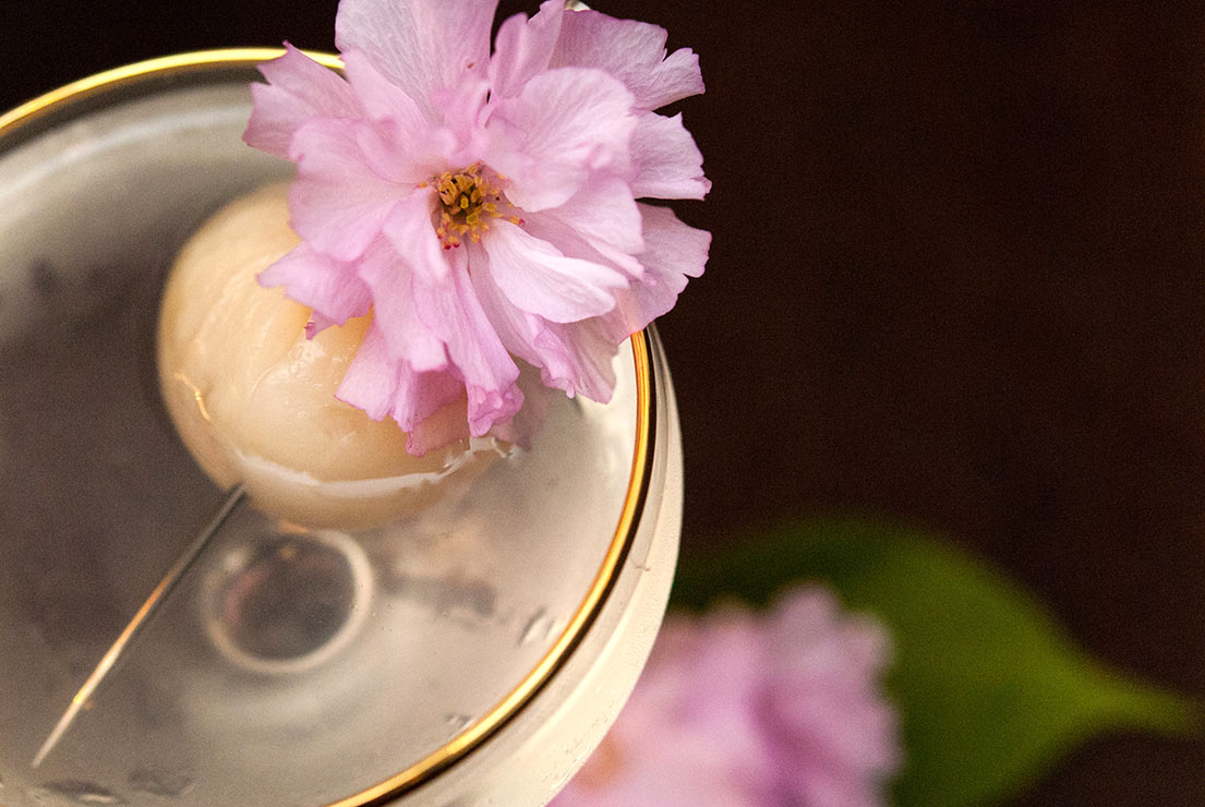 A closeup of a lychee cocktail garnished with a lychee and cherry blossom.