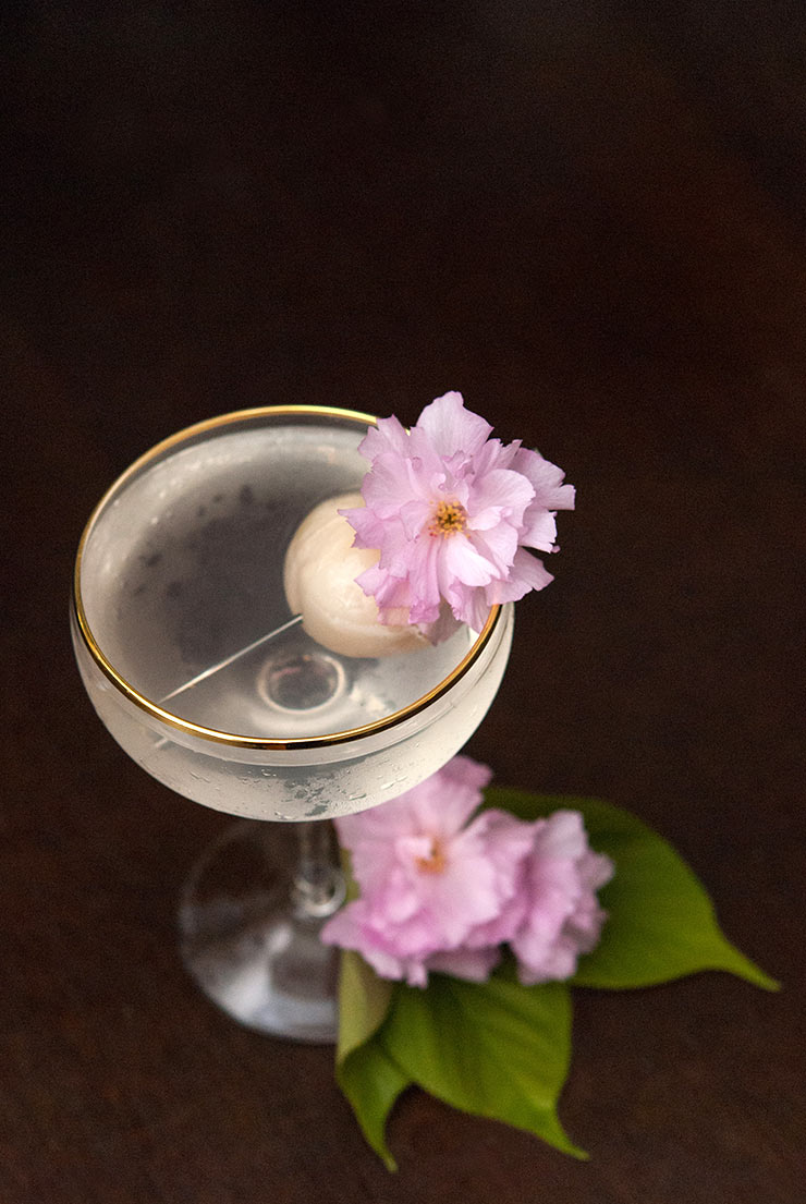 A Lychee cocktail on a dark table, garnished with a lychee and cherry blossoms.