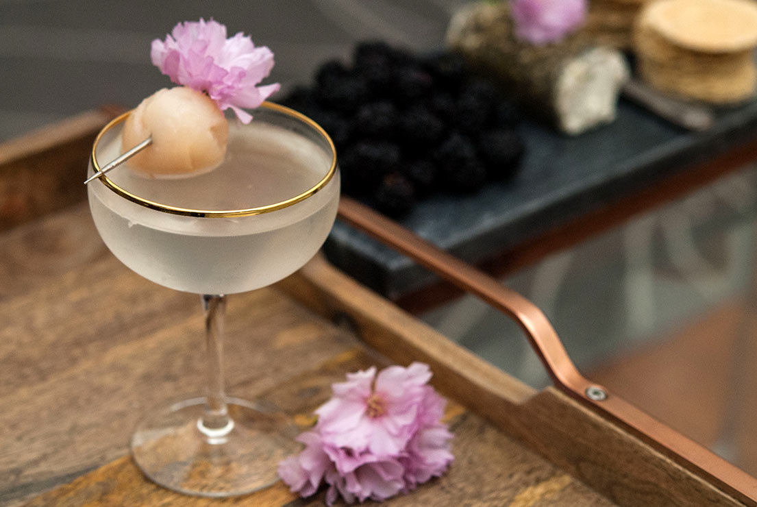 A lychee cocktail garnished with cherry blossoms and a lychee on a wooden tray in front of a cheese plate.