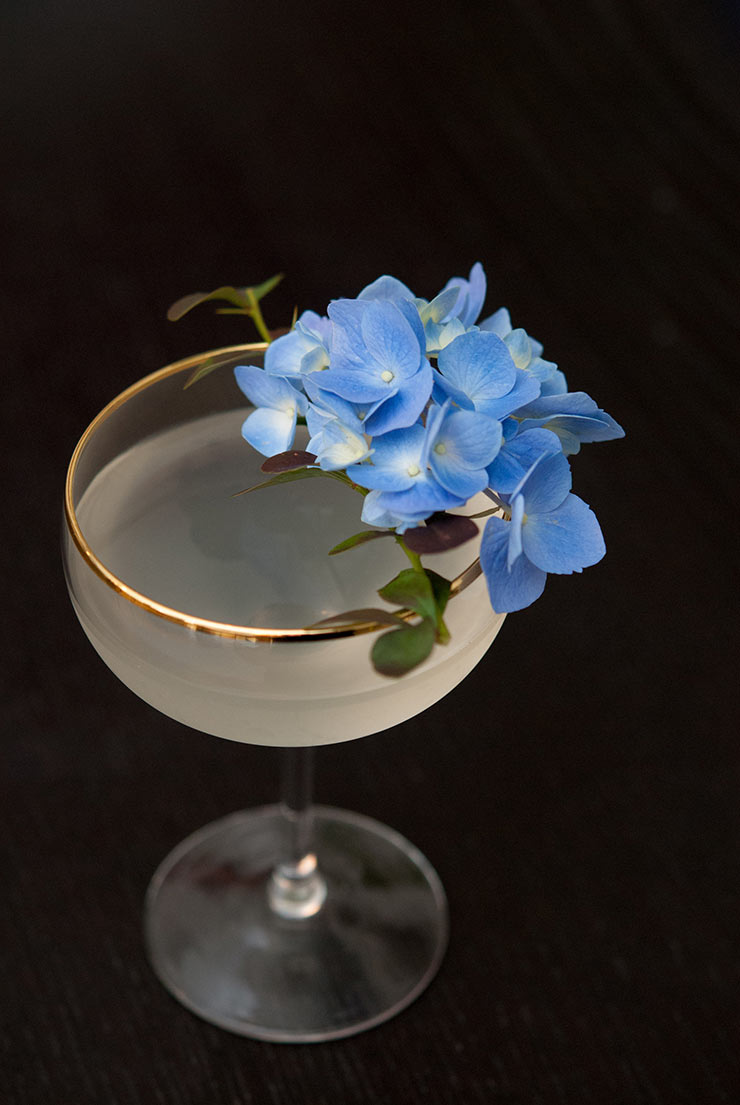 A cocktail with a gold rim, garnished with hydrangea flowers on a black table.