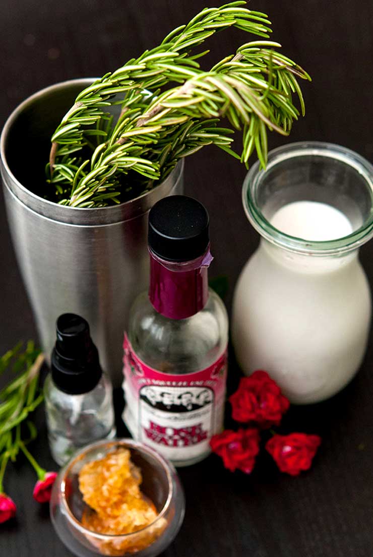 Rosemary in a cocktail shaker, a small bottle of milk, rosewater, honey and spray bottle on a table with a few roses.