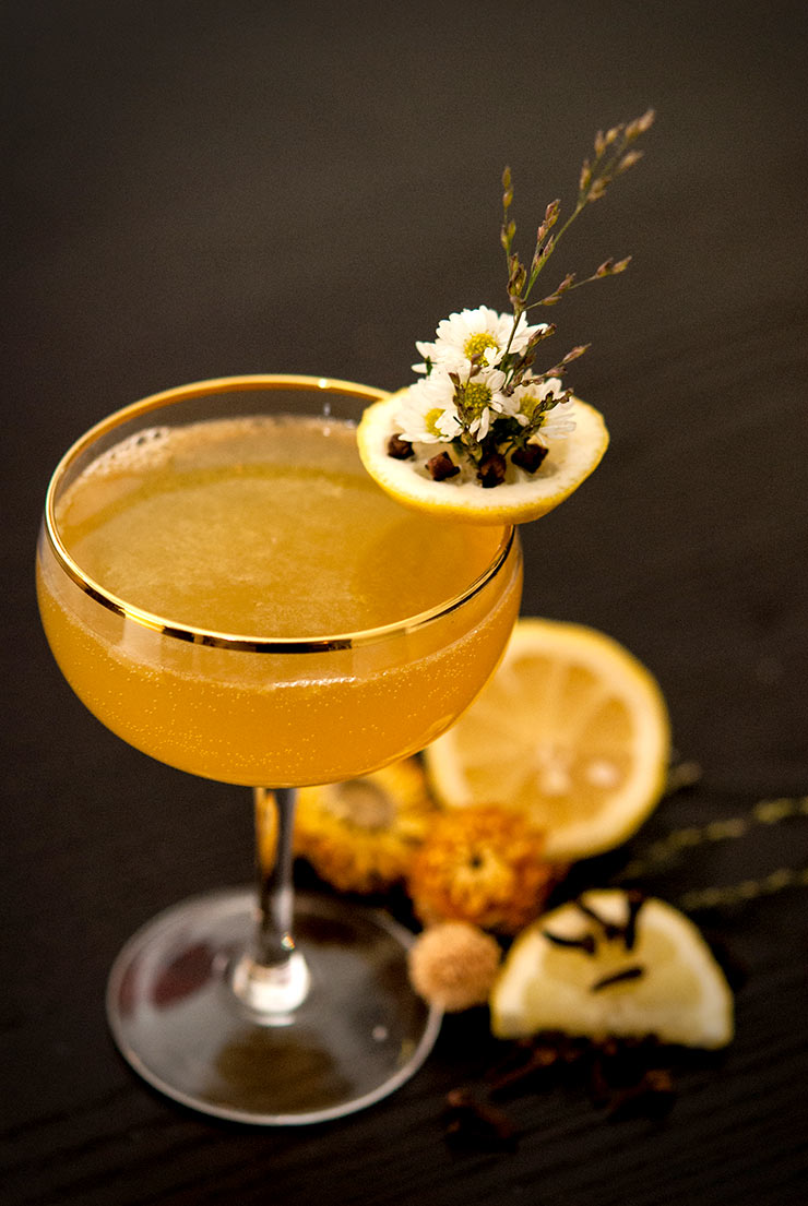 A cocktail garnished with daisies, lemon and cloves, sitting on a black table with sliced lemons and dry flowers around it.